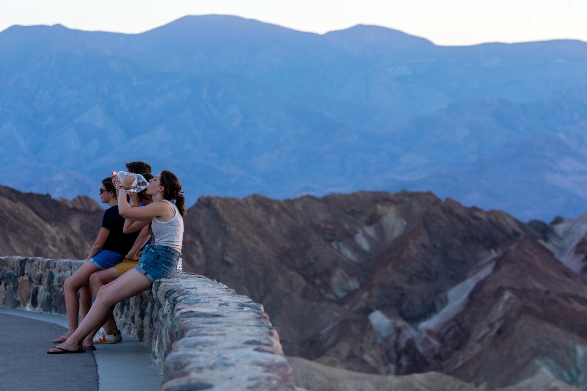 Three people sit on a short stone wall, one drinking water, while an arid mountain range fills the horizon. 