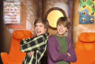 Dylan Sprouse in a flannel shirt with his arms crossed leaning against Cole Sprouse in a purple jacket and a green shirt. 