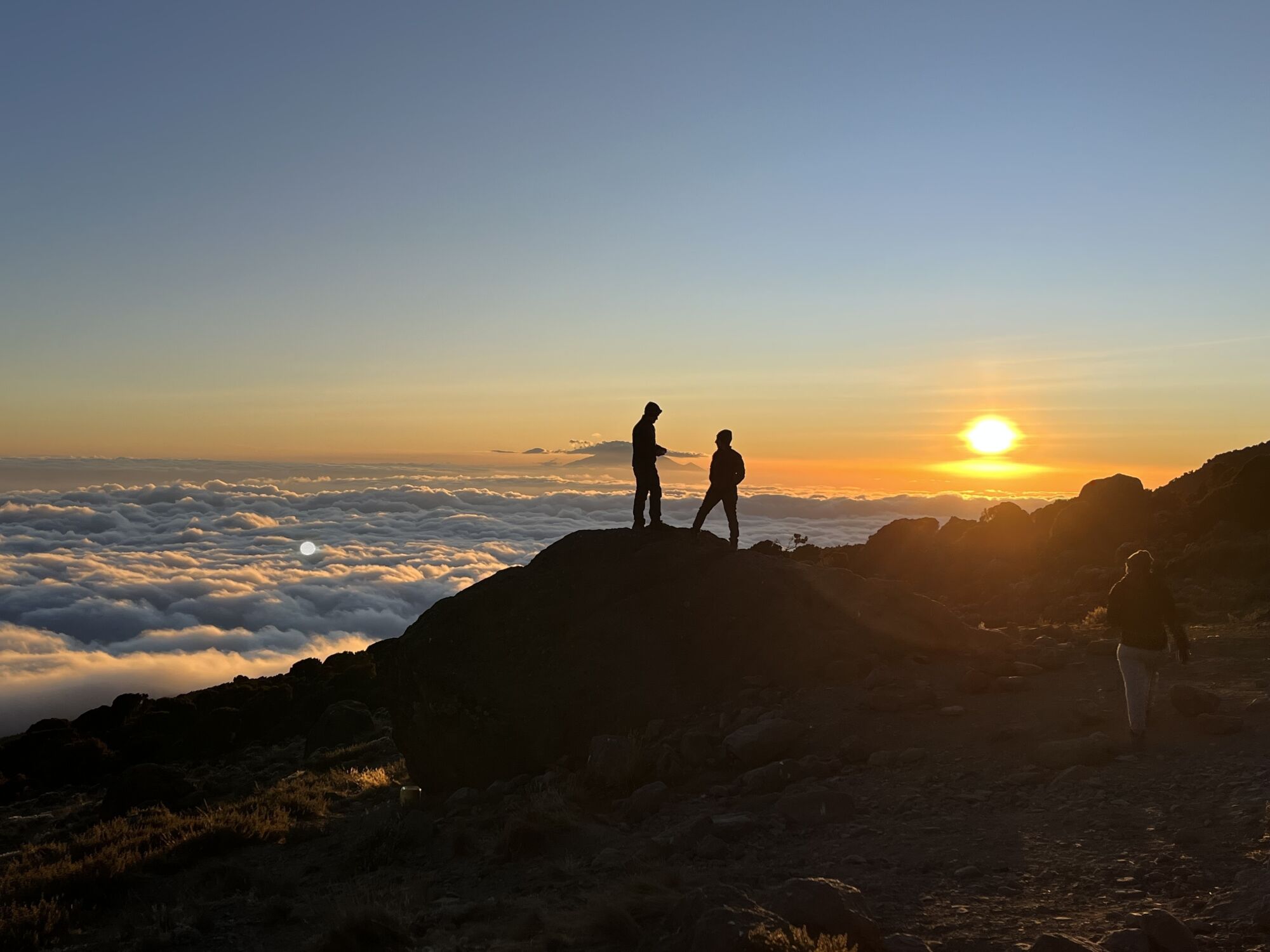 Two people, seen in silhouette in the distance, stand on a peak above clouds as the sun goes down