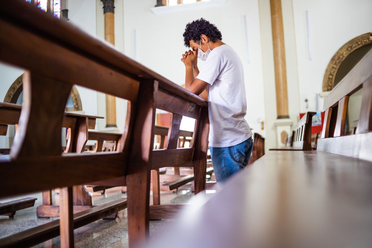 Young man, wearing a white protective mask, inside the church, praying in times of coronavirus