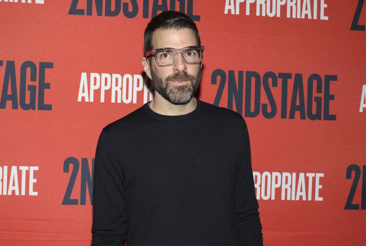 Zachary Quinto standing in a long-sleeve black shirt in front of a red backdrop with black and white lettering