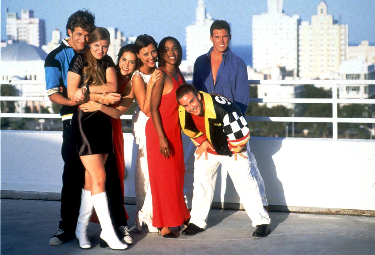 Seven young adults in 1990s attire posing in a group outdoors by a white railing with a cityscape in the background