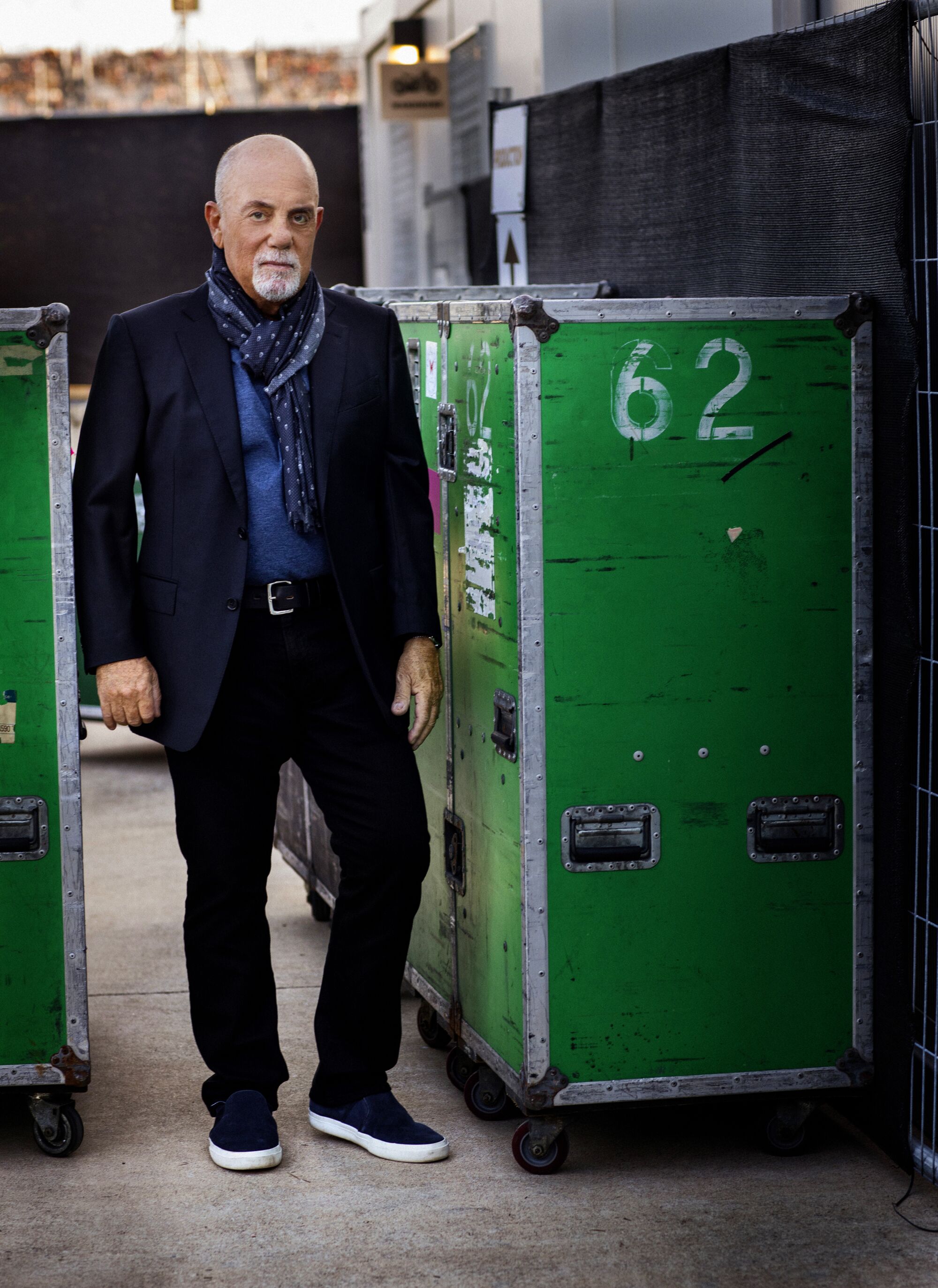 Billy Joel, dressed in dark clothing, stands between large rolling crates.
