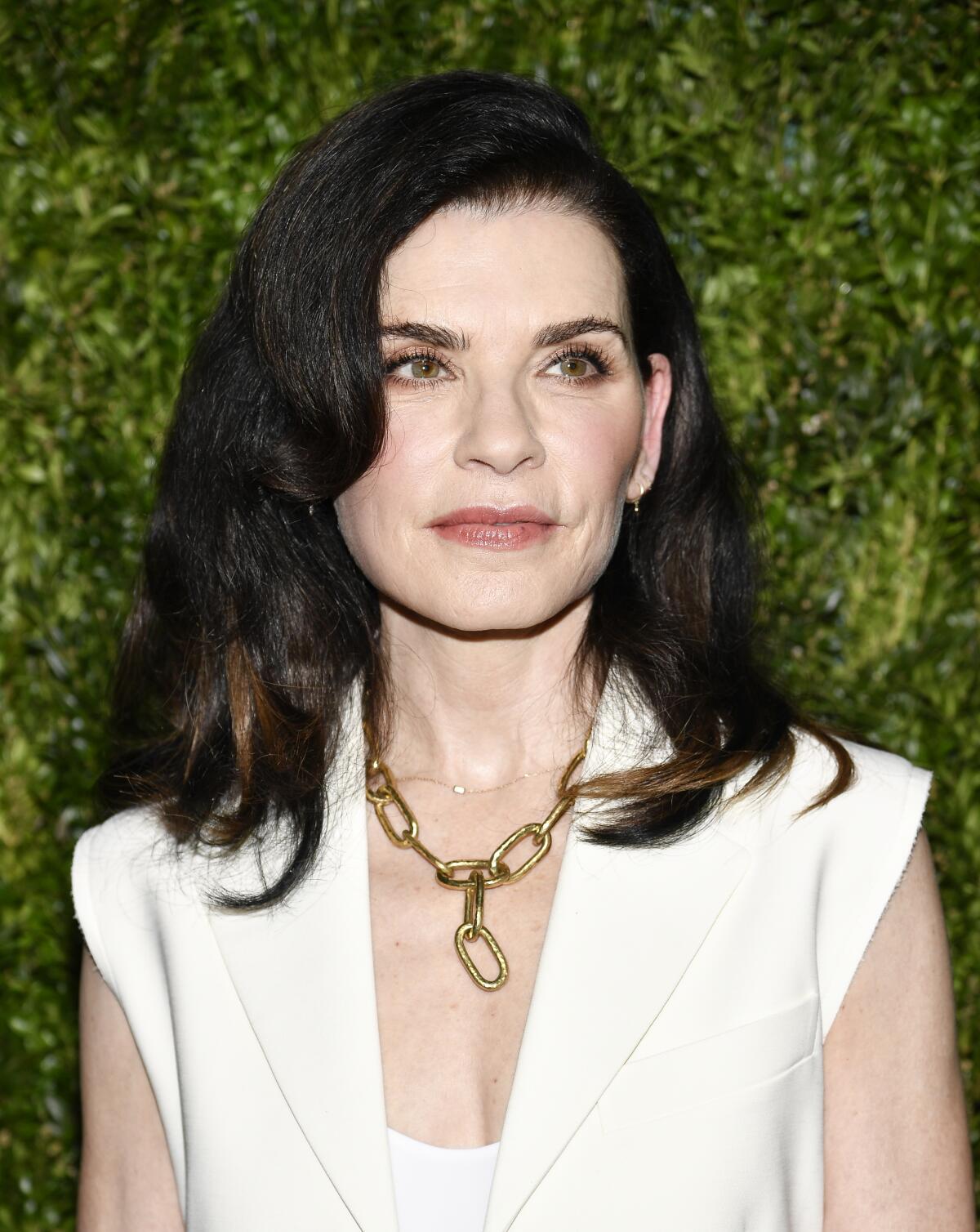 Julianna Margulies wears a cream colored vest over a white blouse with a chunky gold chain necklace.