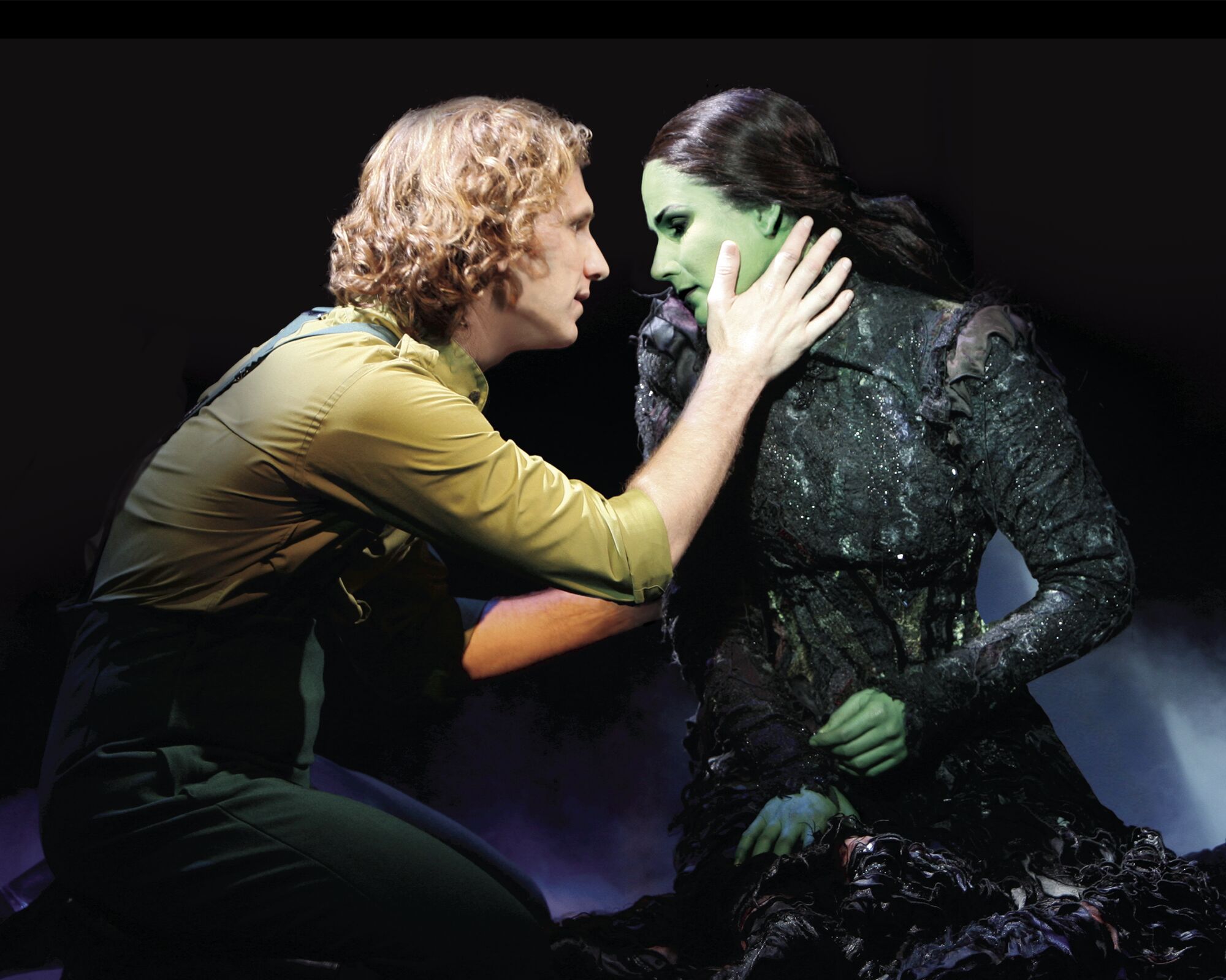 A man kneels next to a woman, putting his hand on her face, which is green