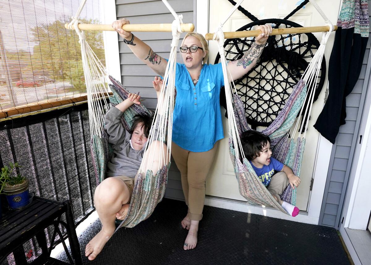 A blonde woman plays with two children in swings on a porch of a home.