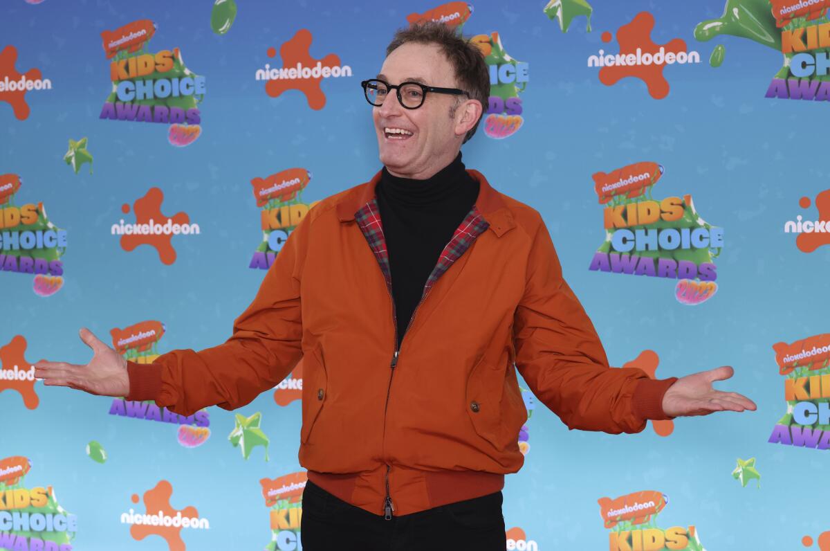 Tom Kenny, in an orange jacket and black turtleneck, stands holding his arms out and palms up
