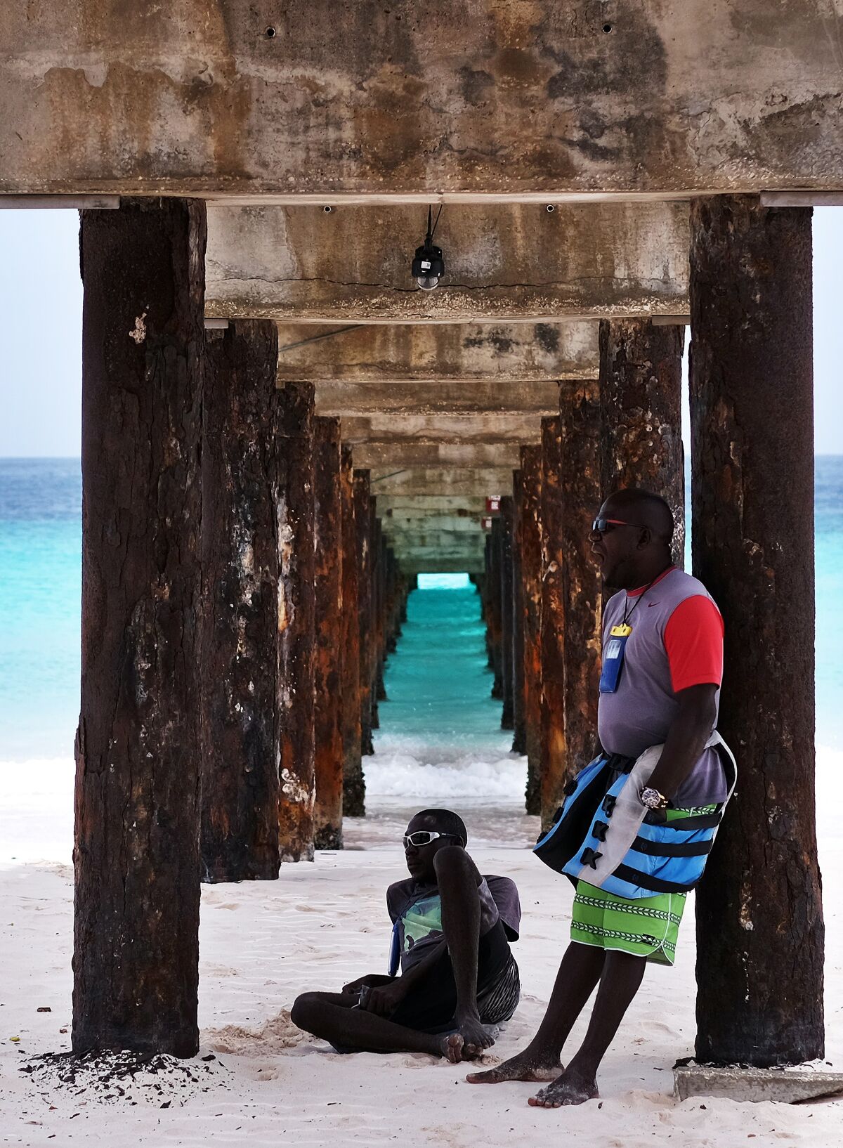 Two people stand under a jetty on a beach in Barbados.