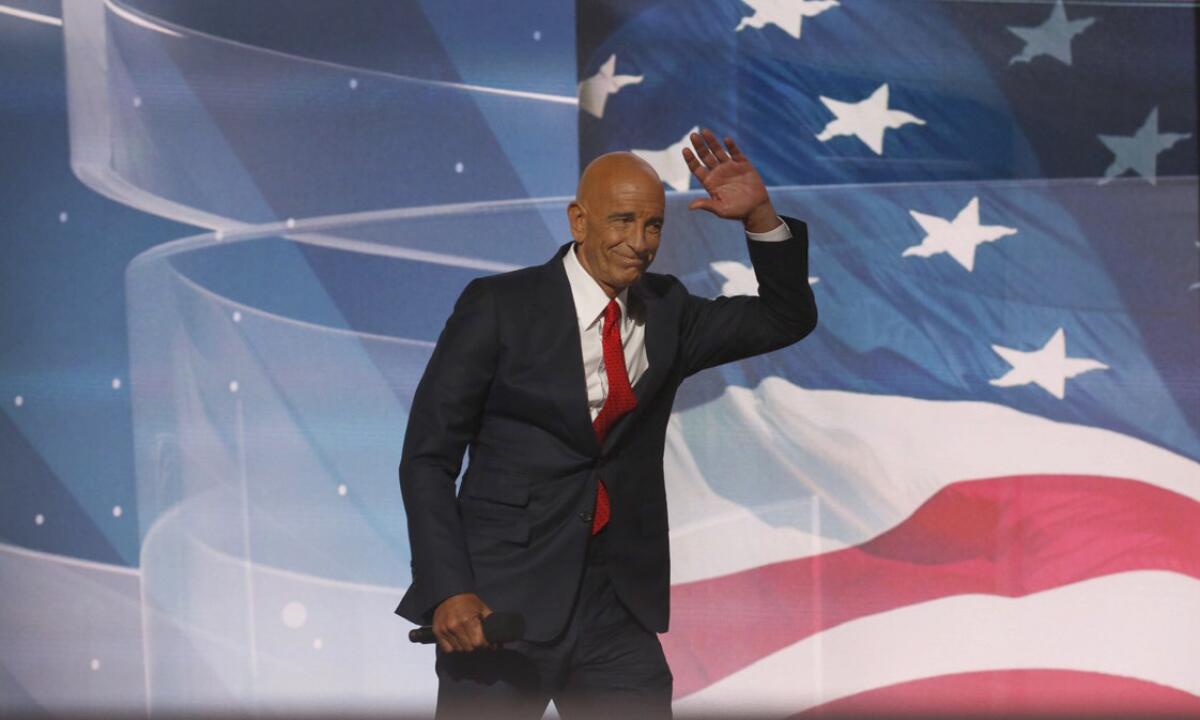 Tom Barrack waves as he walks in front of an image of a U.S. flag