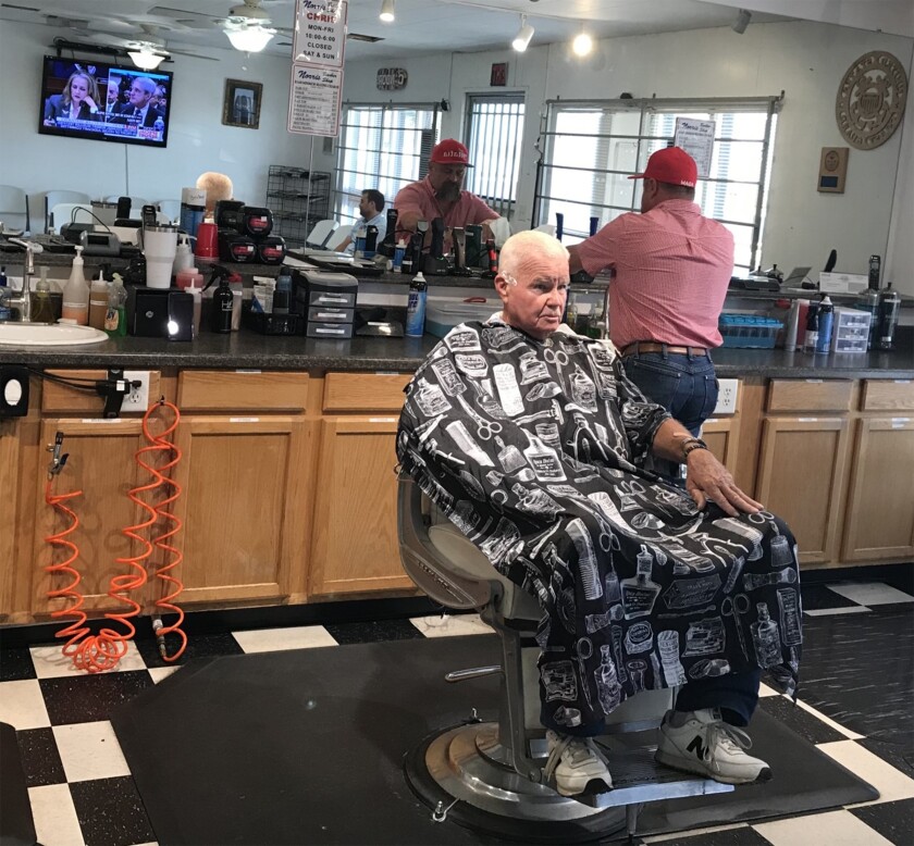 Oildale, Calif., barber Chris Vaughn, in the red Trump hat, finishes up a shave for a regular customer. In the background, the Robert S. Mueller III hearings are on TV.