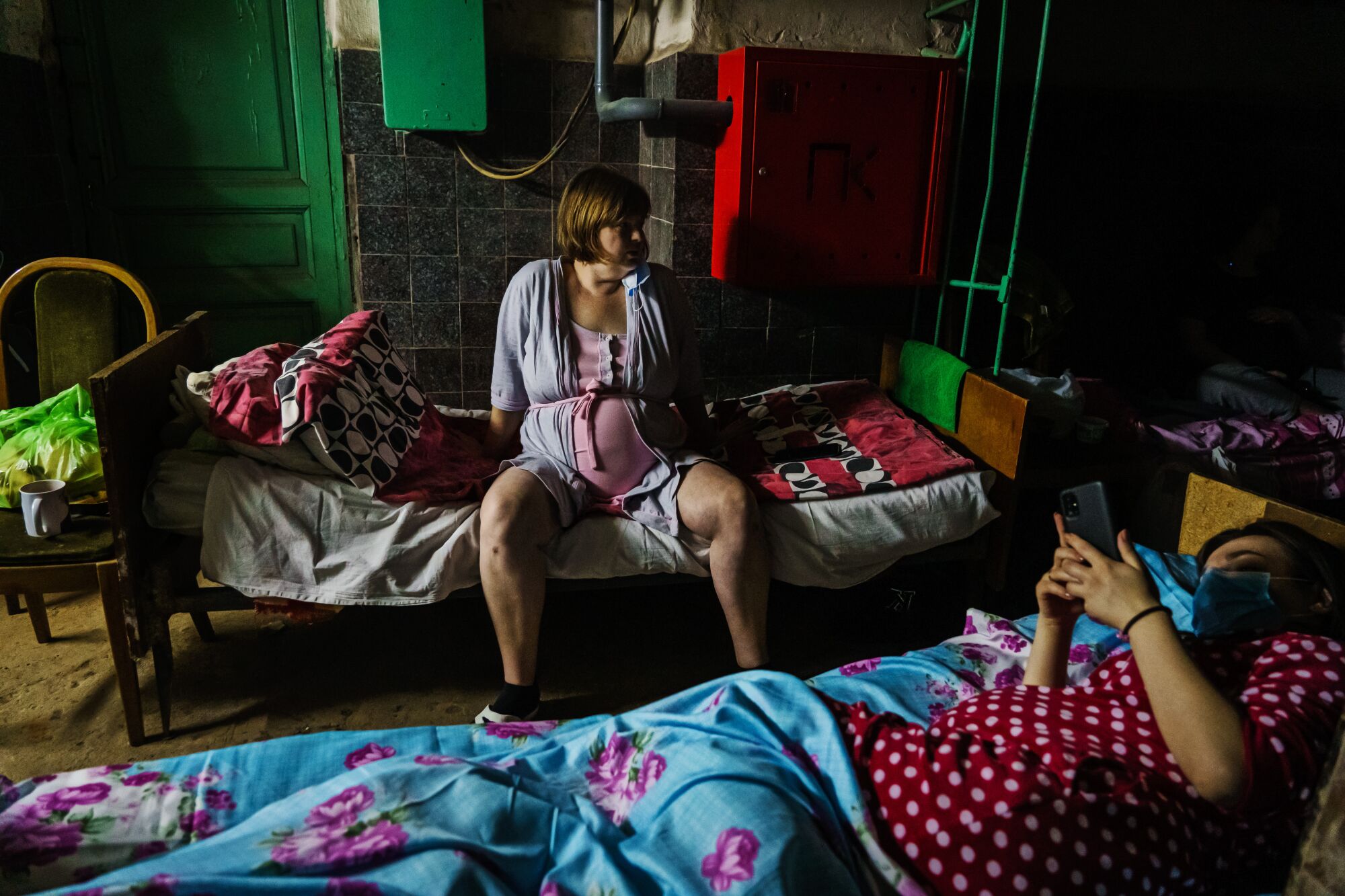 Pregnant women, one sitting up and one lying down looking at her phone, on beds in a dark shelter.