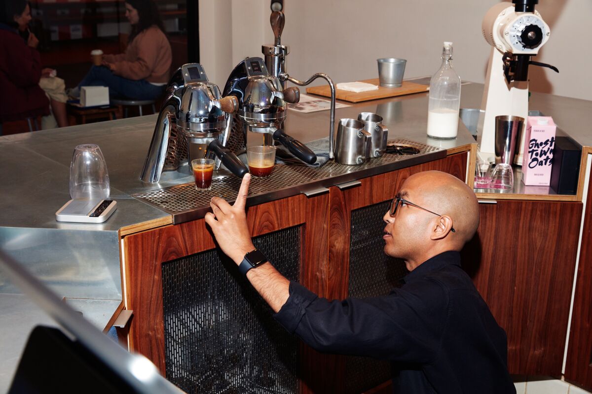 Behind a service counter, a man kneels to view the viscosity of espresso in a clear glass.