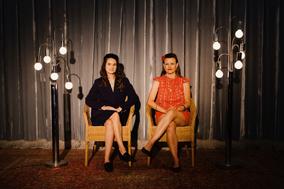 Two women sit in chairs between lights and in front of a curtain.
