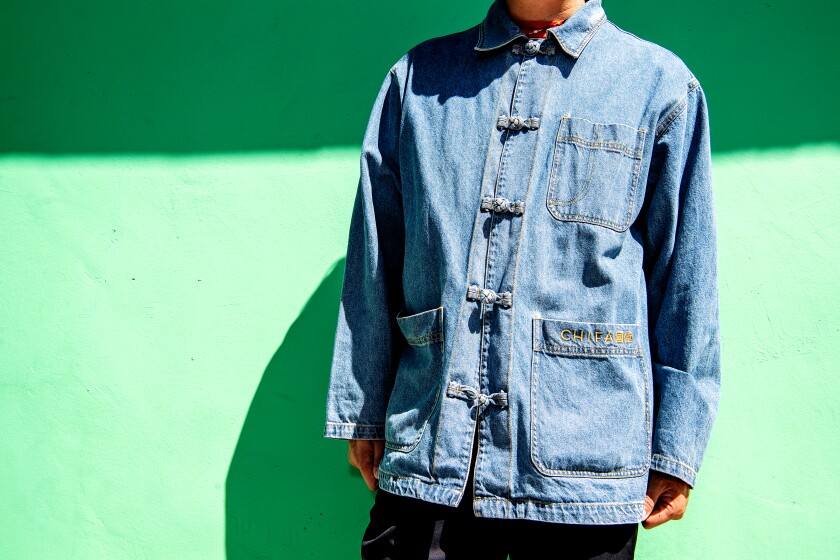 A man in a denim smock stands against a green wall.