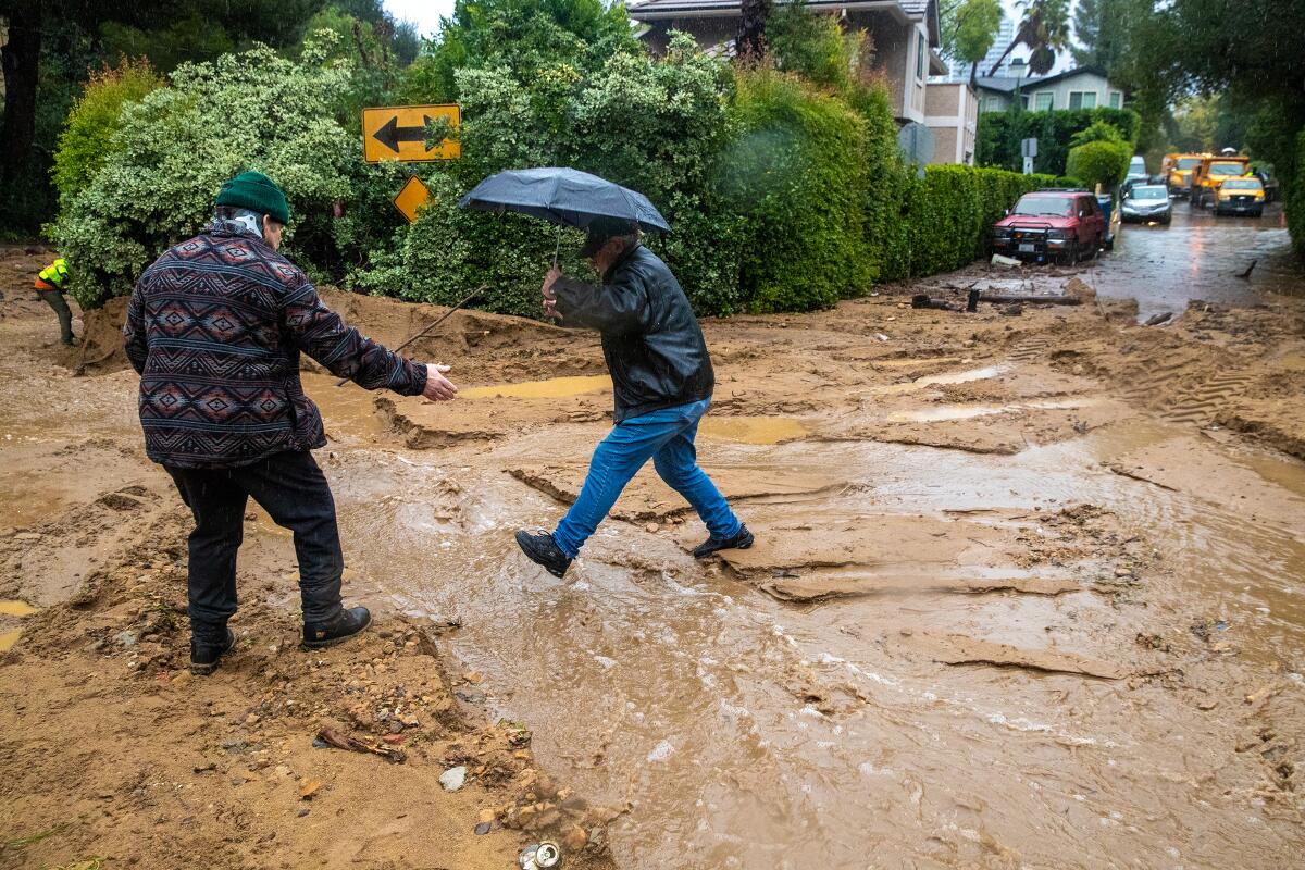 A man, left, extends a hand toward another man holding an umbrella as he steps across a flow of water on a muddy street