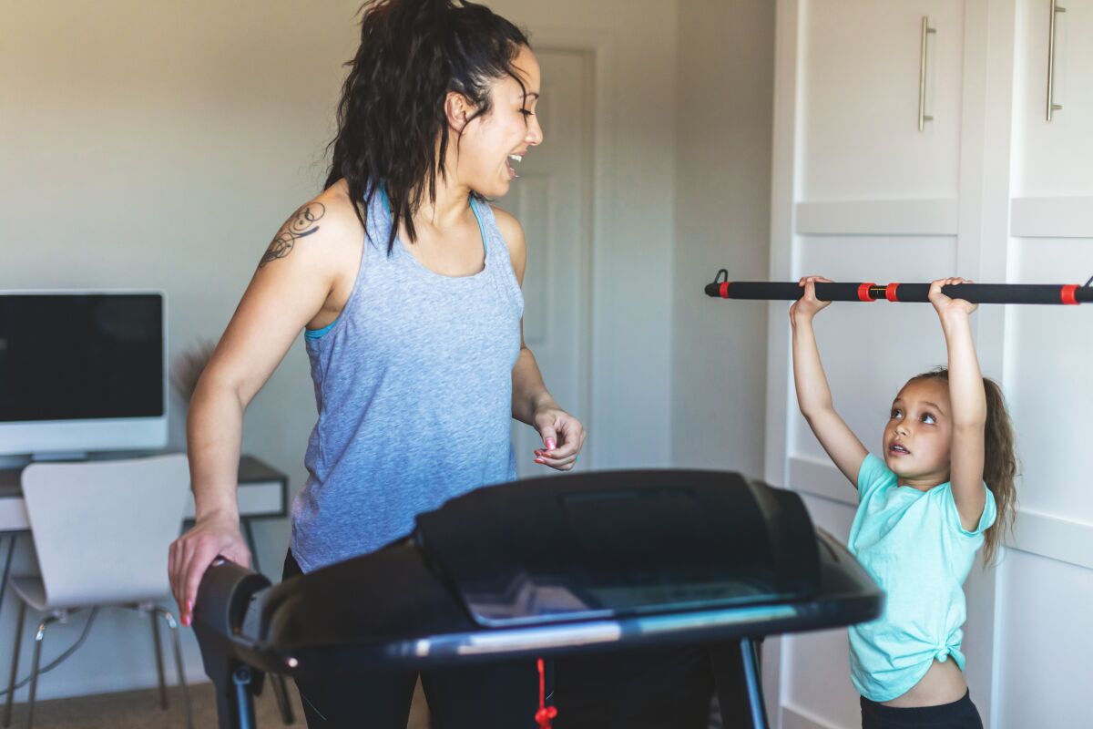 A mom exercises on a treadmill while her young daughter stands next to her on the floor, holding an empty weight bar.