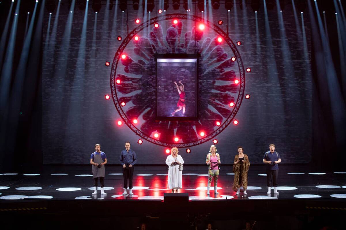Six performers are seen on stage while another is suspended in a circular structure above in "Covid fan Tutte"