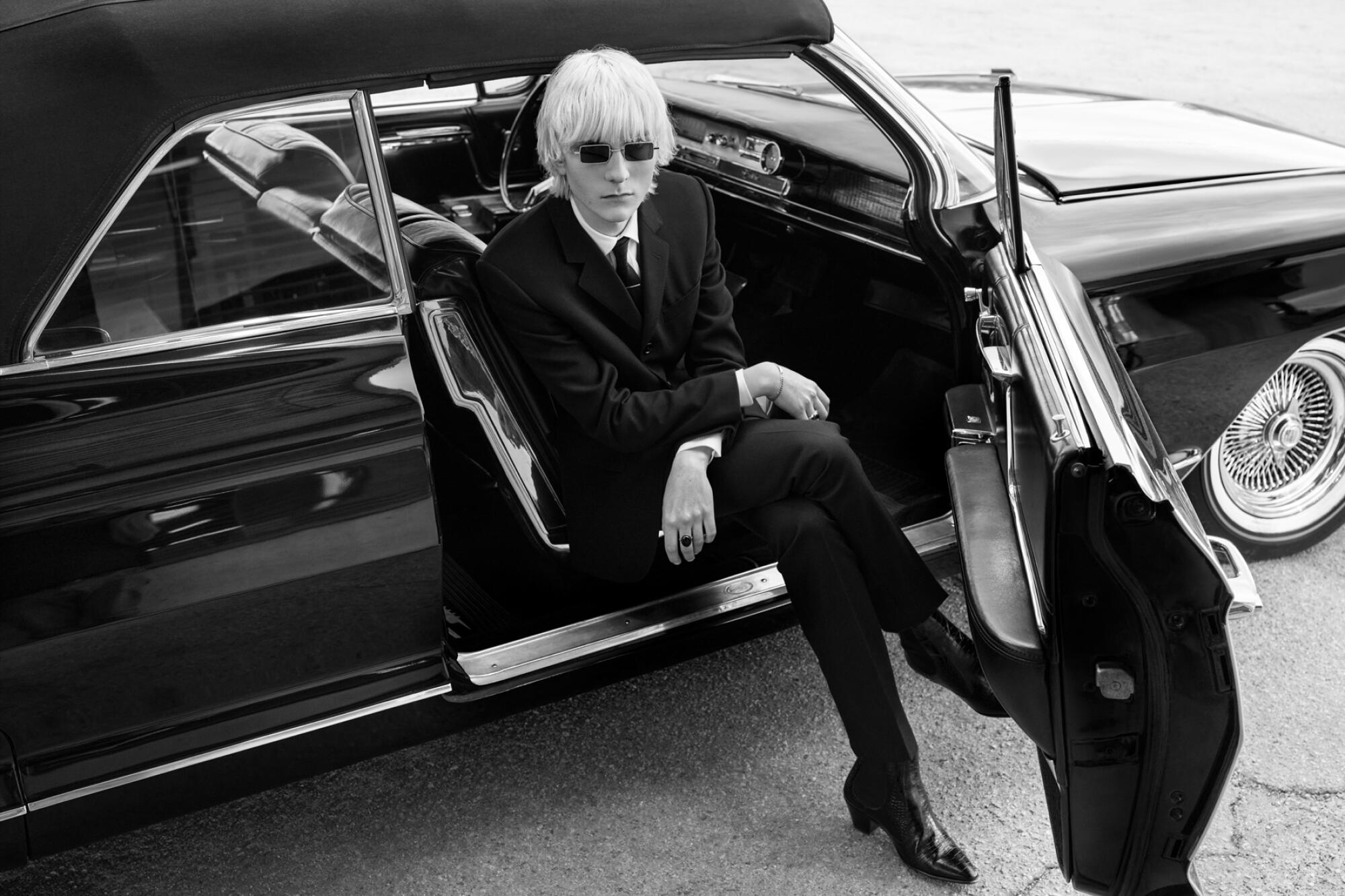 A black-and-white vintage-looking photo of a model seated in a car, its door open