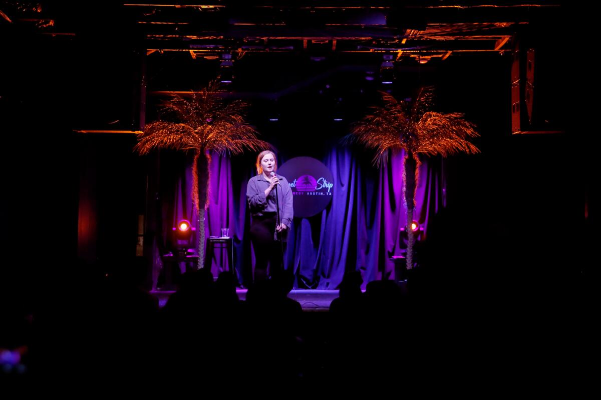 Woman standing onstage, speaking into a microphone, between two palm trees