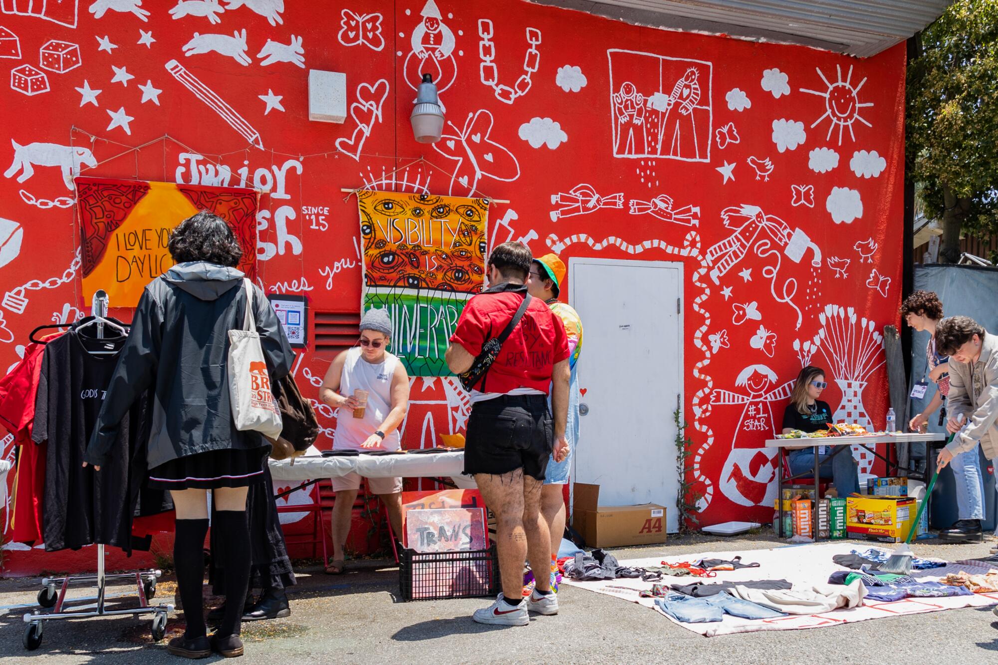 People standing outside a building that has a bright red mural, with a couple of vendors with tables. 