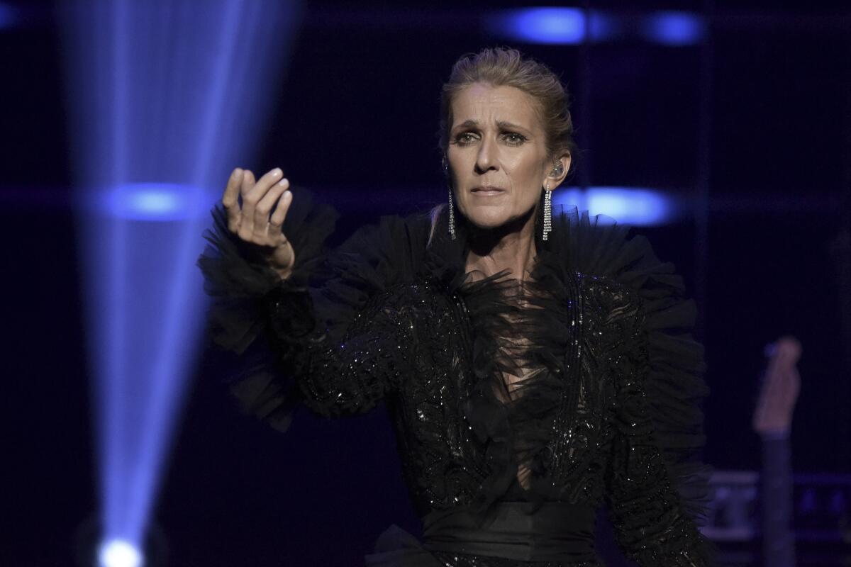 Celine Dion standing on a stage backlit with blue lights in a black suit holding her arm in front of her.