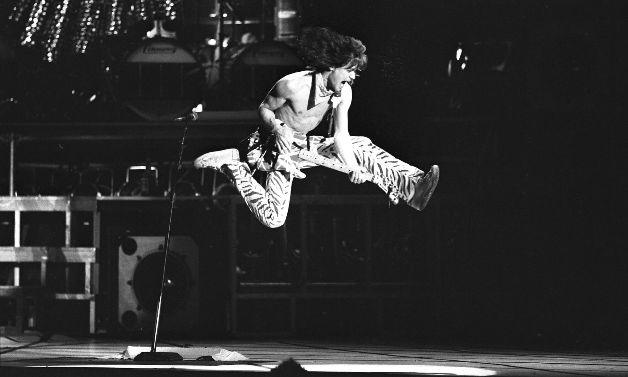 Eddie Van Halen, shirtless and with a bandanna around his neck, leaps in the air as he plays his guitar.