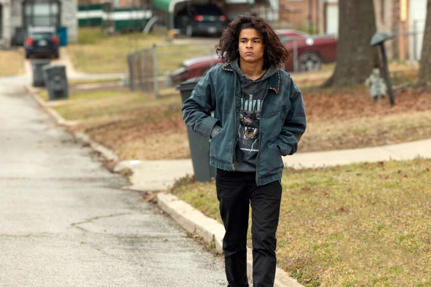 A long-haired teenager in a denim jacket is walking down a street on a gray day.