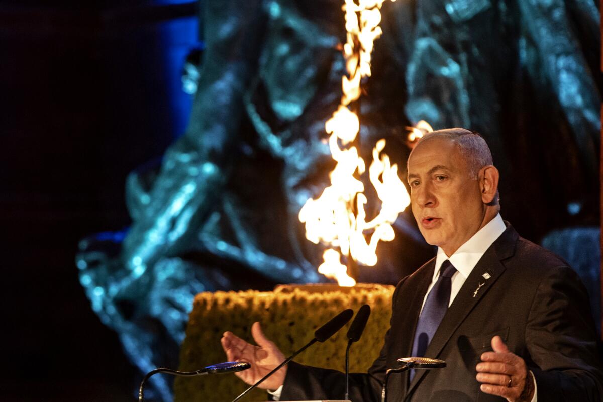 Benjamin Netanyahu speaks into microphones, with a flame behind him. 