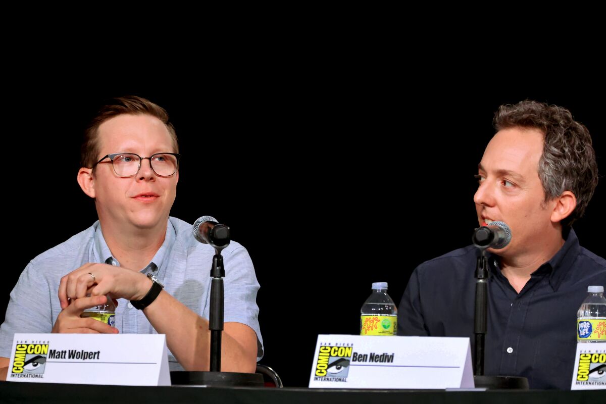 Two men sit behind microphones at a table against a black background. Place cards show their names.
