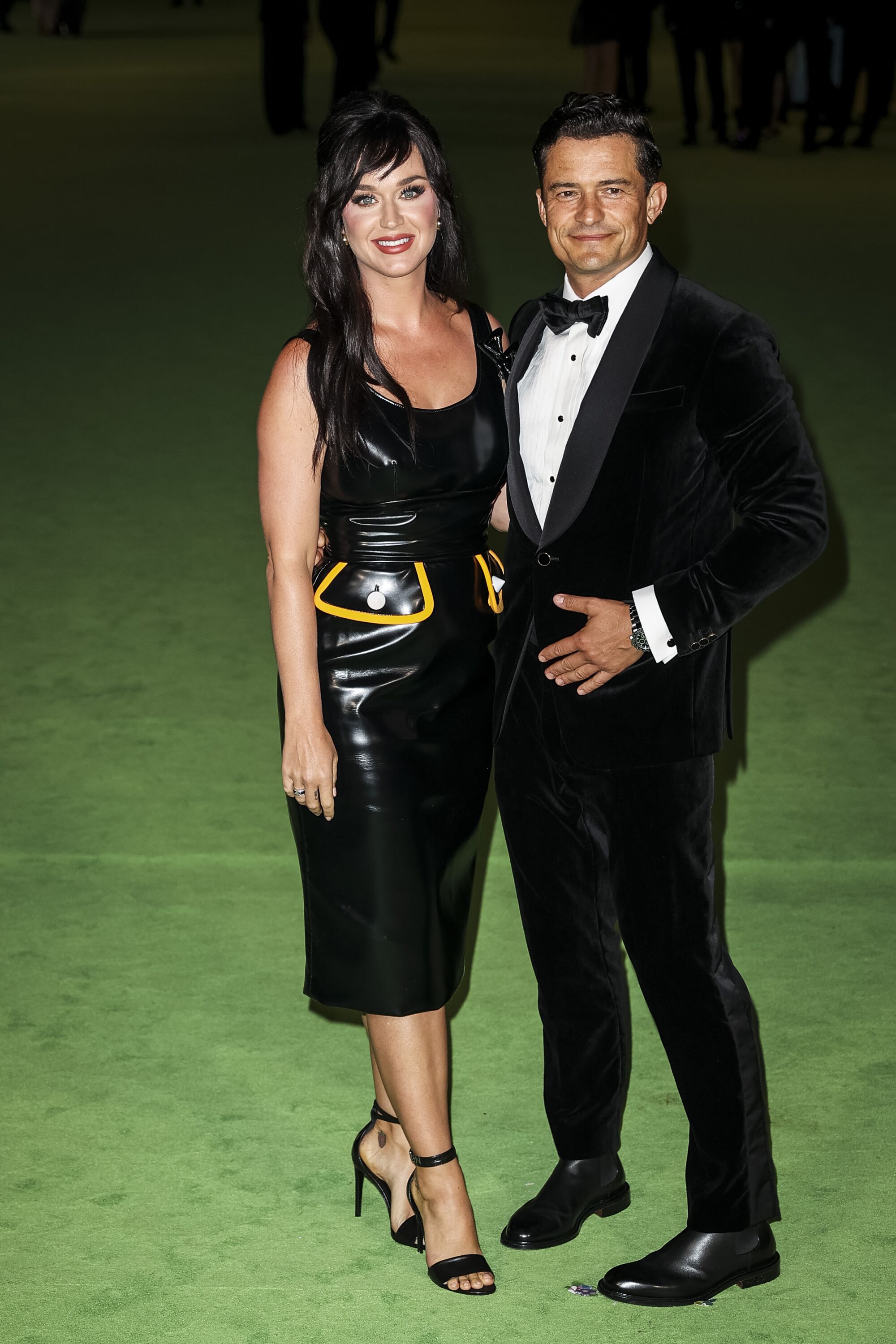 A woman in a black dress and a man in a black tuxedo posing on a green carpet
