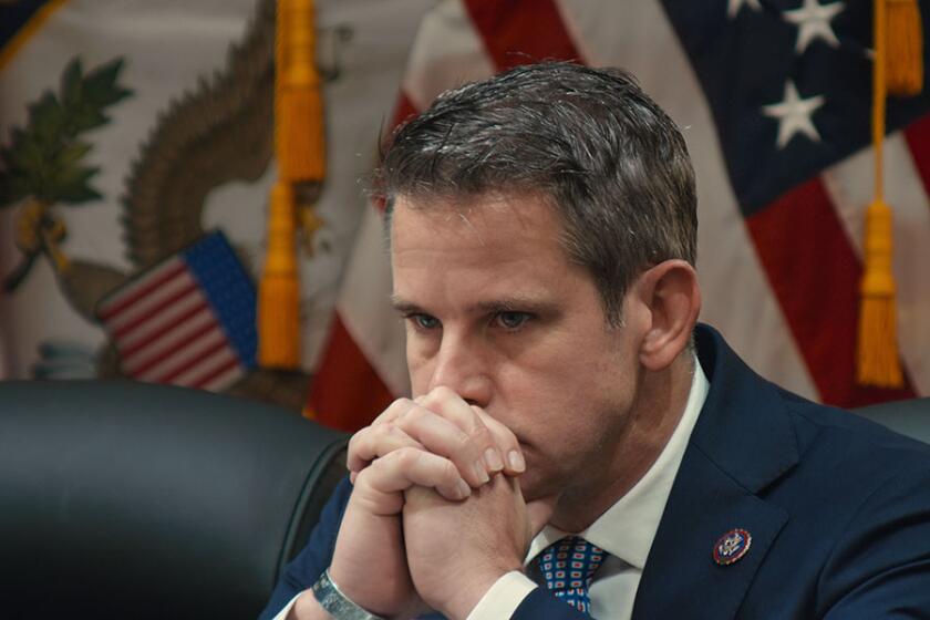 A congressman with flags behind him holds his hands to his face.