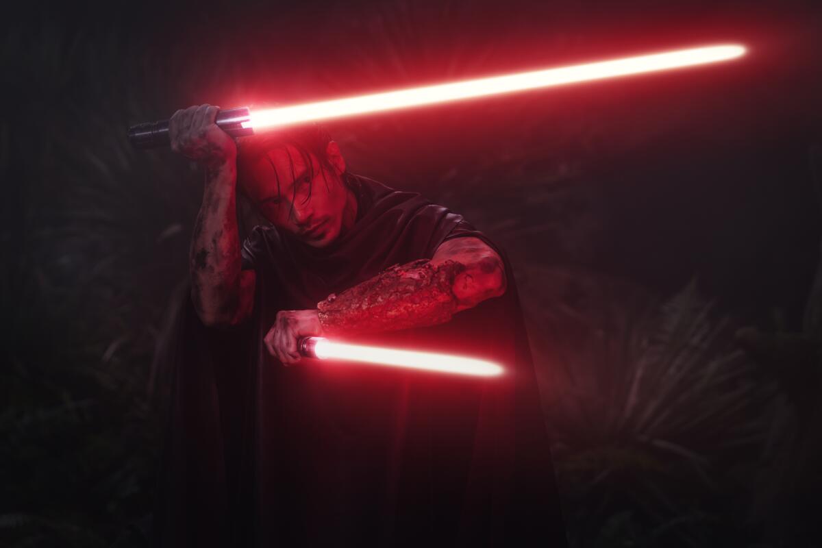 Manny Jacinto, in a cloak, wields two red lightsabers.