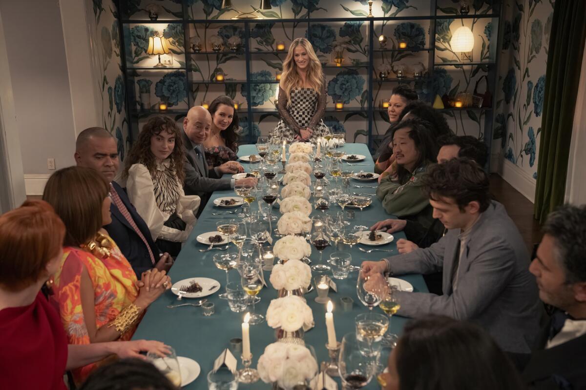 Carrie Bradshaw at the head of a long dinner table covered in a blue tablecloth, with people seated on each side.