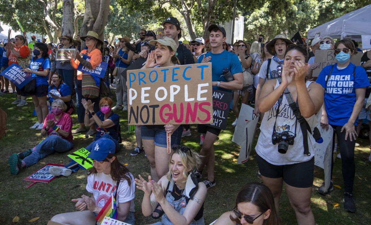 A crowd of demonstrators standing and sitting, one holding a handmade sign reading, "Protect people, not guns."