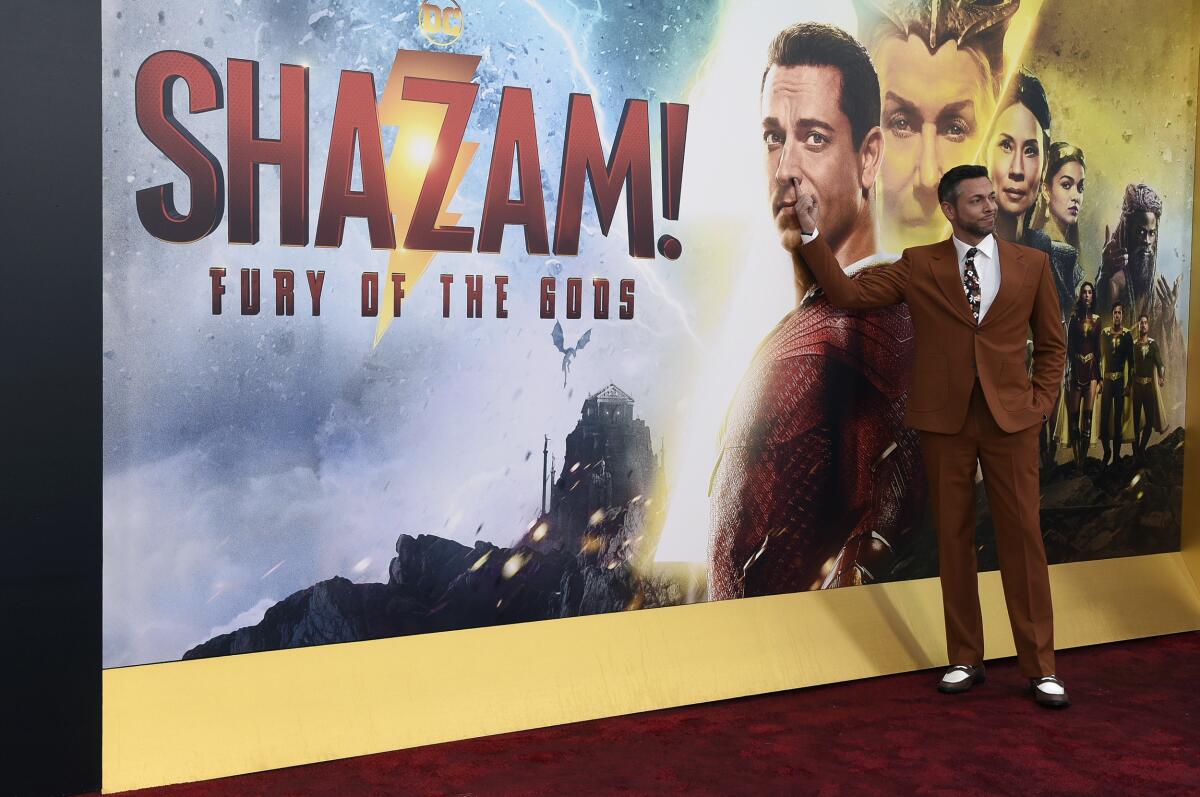 A man in a suit stands on a red carpet in front of a huge movie-poster background