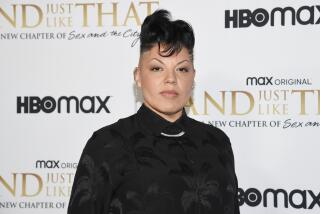 Sara Ramirez in a dark formal shirt and a chain necklace posing in front of a white backdrop with black and gold text