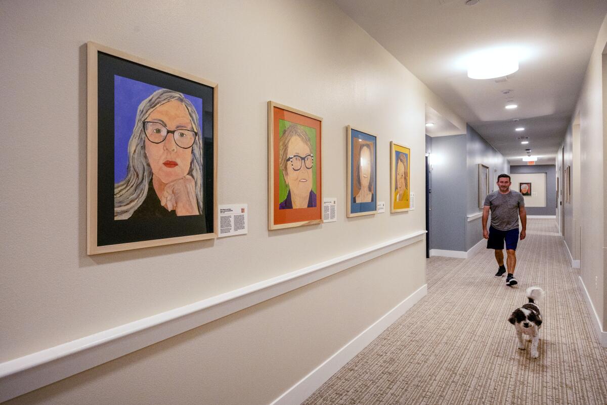 A man and a dog walk in a hallway where "Feeling Our Age"portraits hang at Watermark Laguna Niguel.