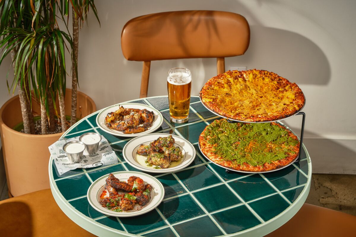 Two pizzas, chicken wings and a glass of beer on a green-tile tabletop.