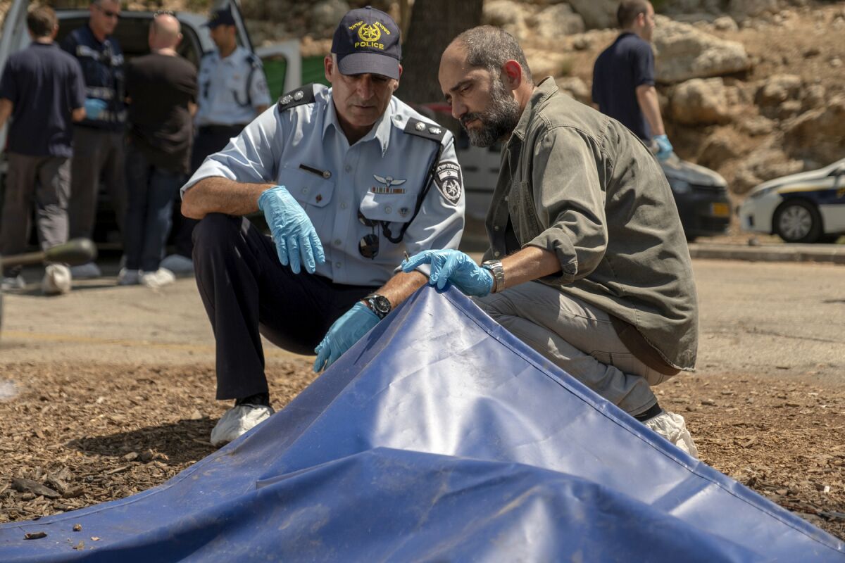 Two men wearing gloves crouch to look under a tarp.