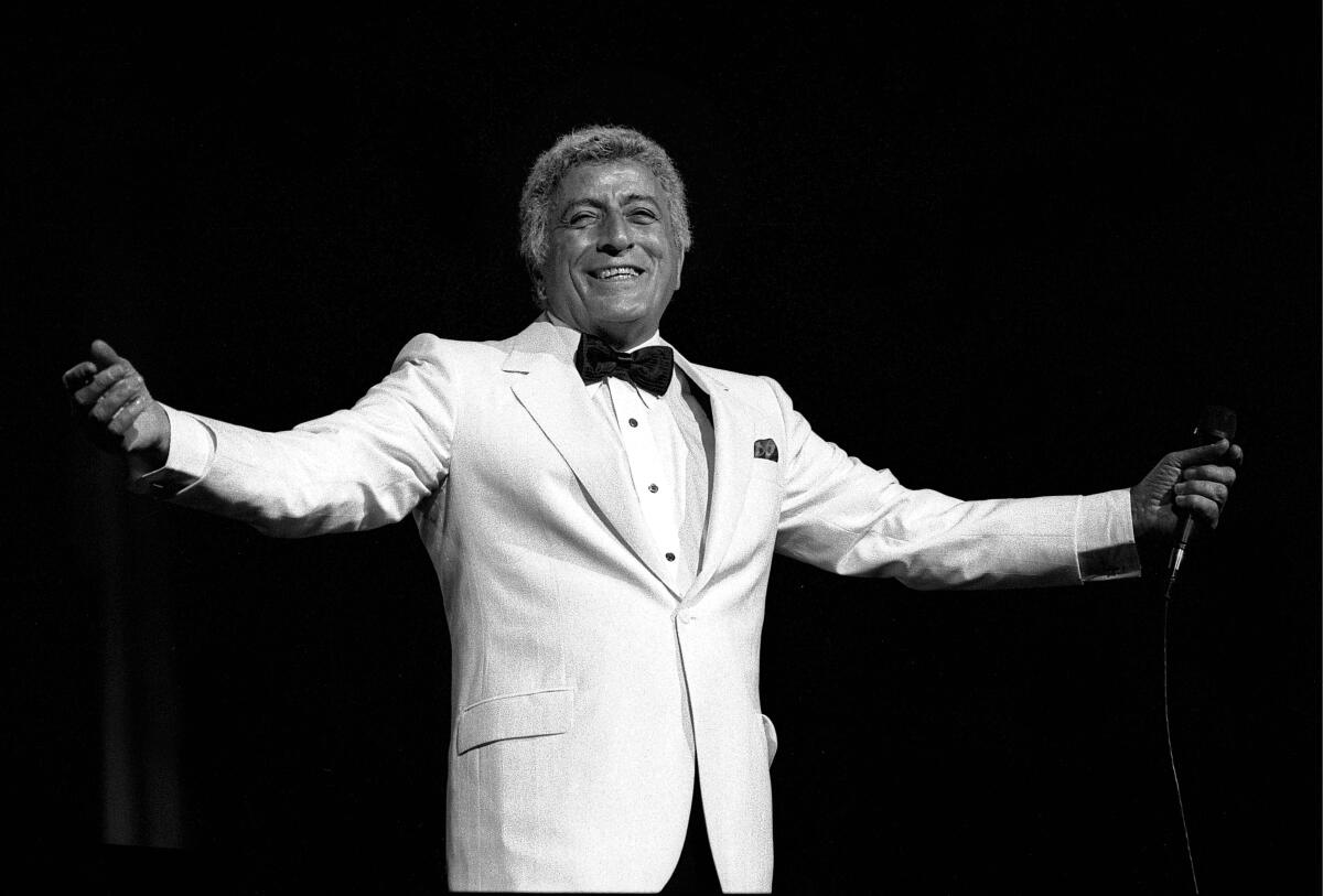 Tony Bennett in a white jacket and black bow tie gestures with outstretched arms.