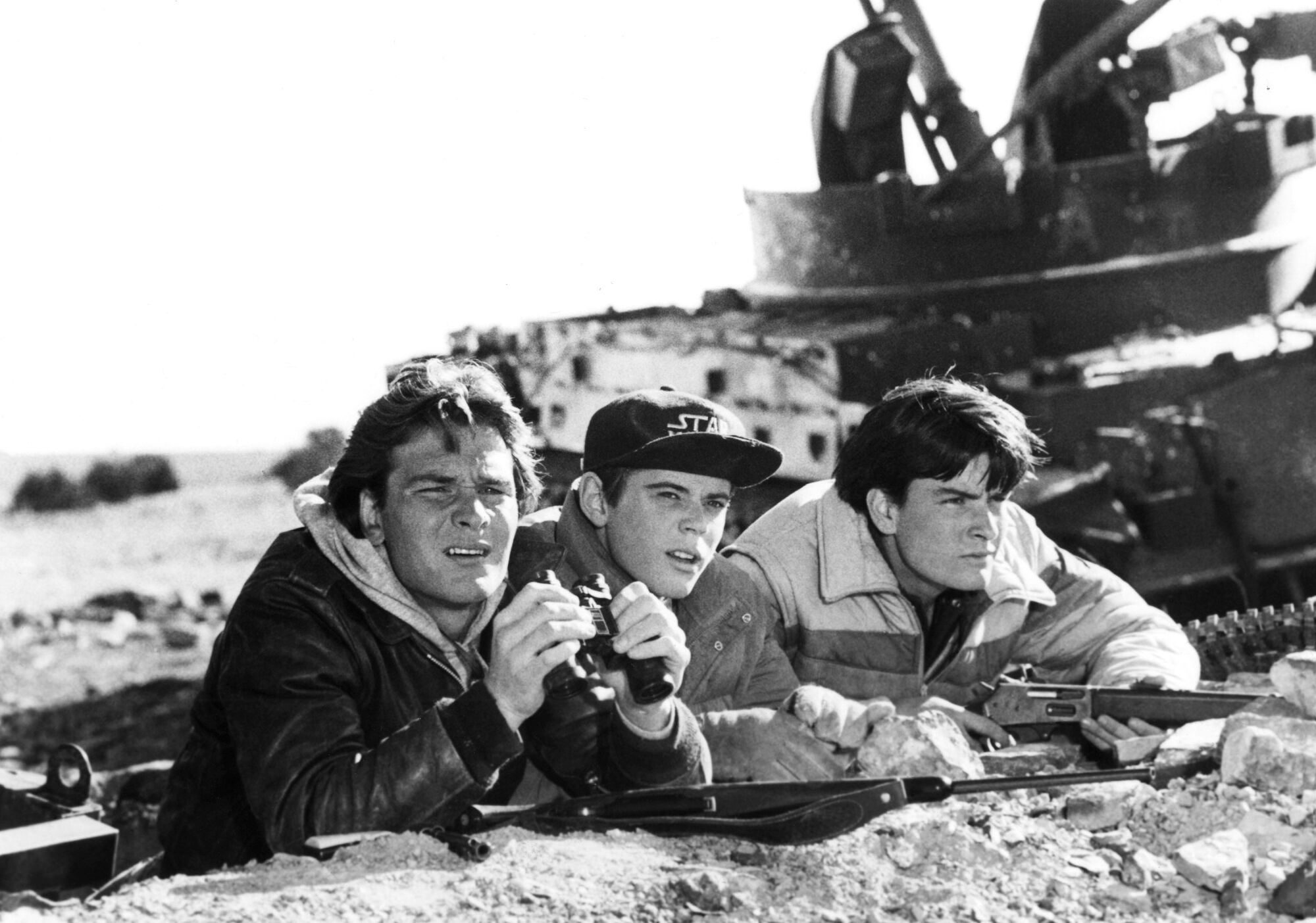 A black-and-white film still shows Patrick Swayze, C. Thomas Howell and Charlie Sheen crouching in front of a tank.