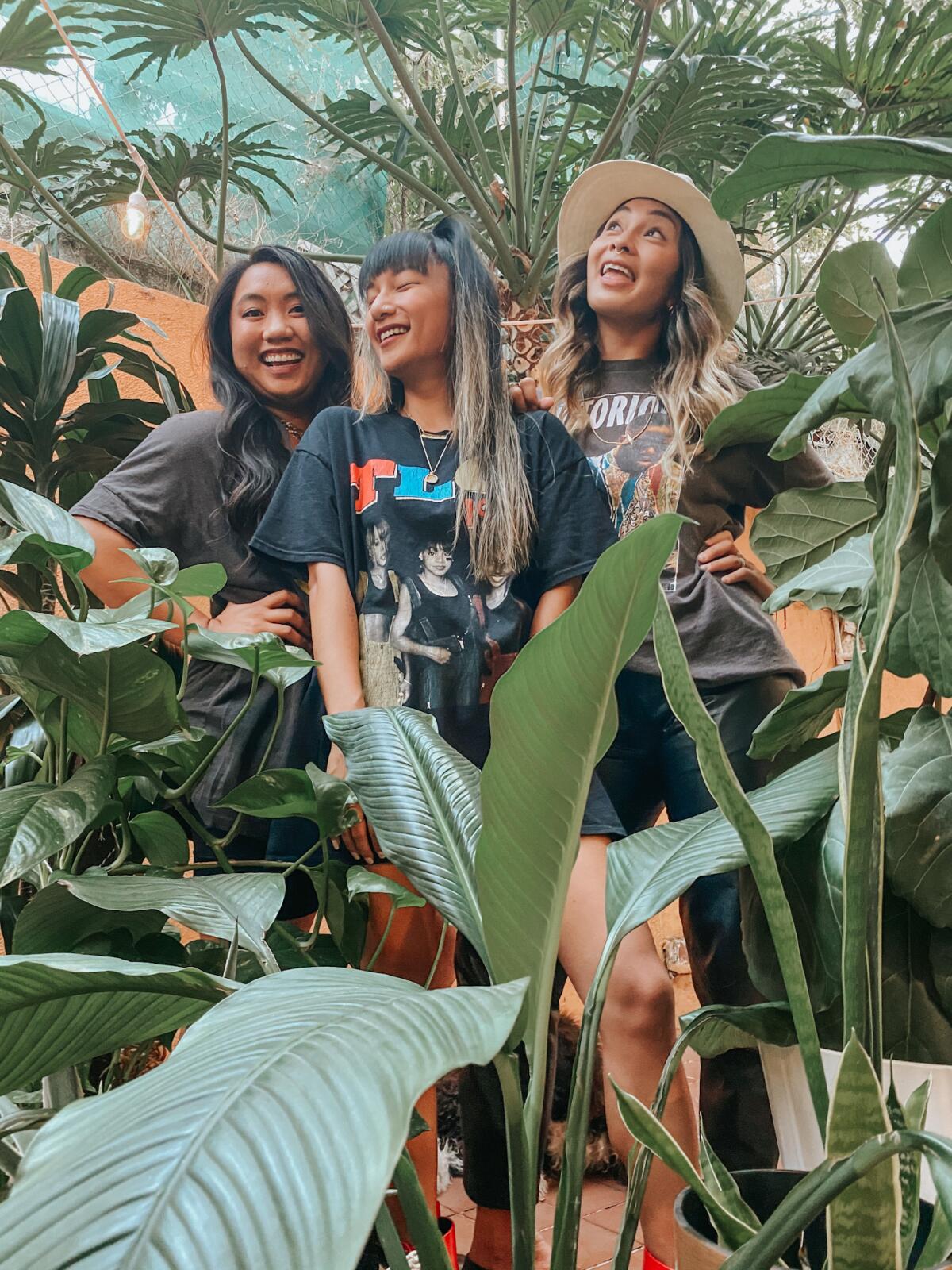 Three women dressed in dark-colored, loose T-shirts smile surrounded by plants and greenery.