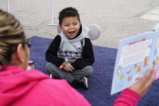 A woman and a little boy sit on a blue carpet while the woman reads a picture book to him.