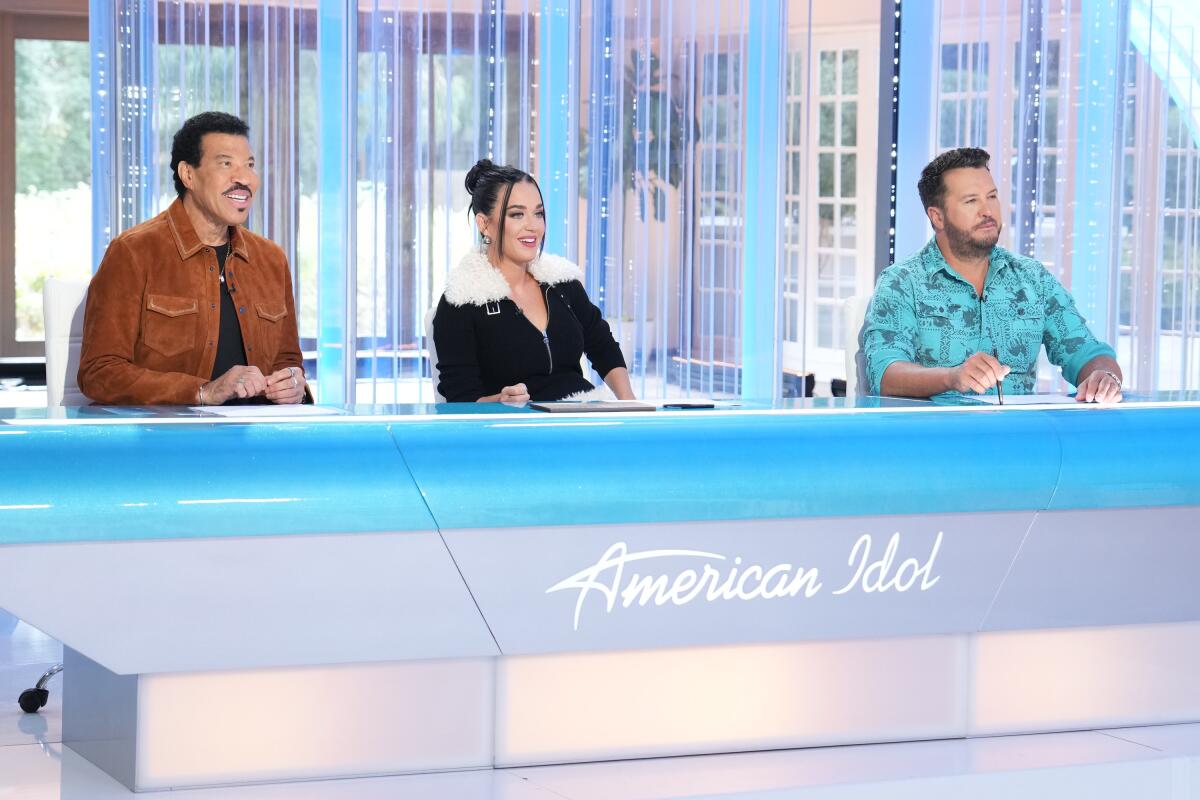 Lionel Richie, Katie Perry and Luke Bryan sitting behind a long blue desk that says "American Idol." 