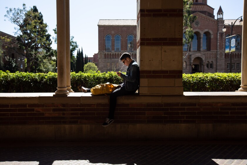 A young person sits between columns on a university campus next to a backpack and looking at a phone.