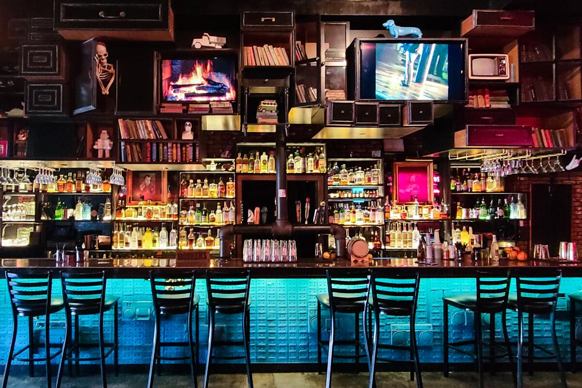 A bar with a large wall of drinks and TV screens and a line of empty barstools 