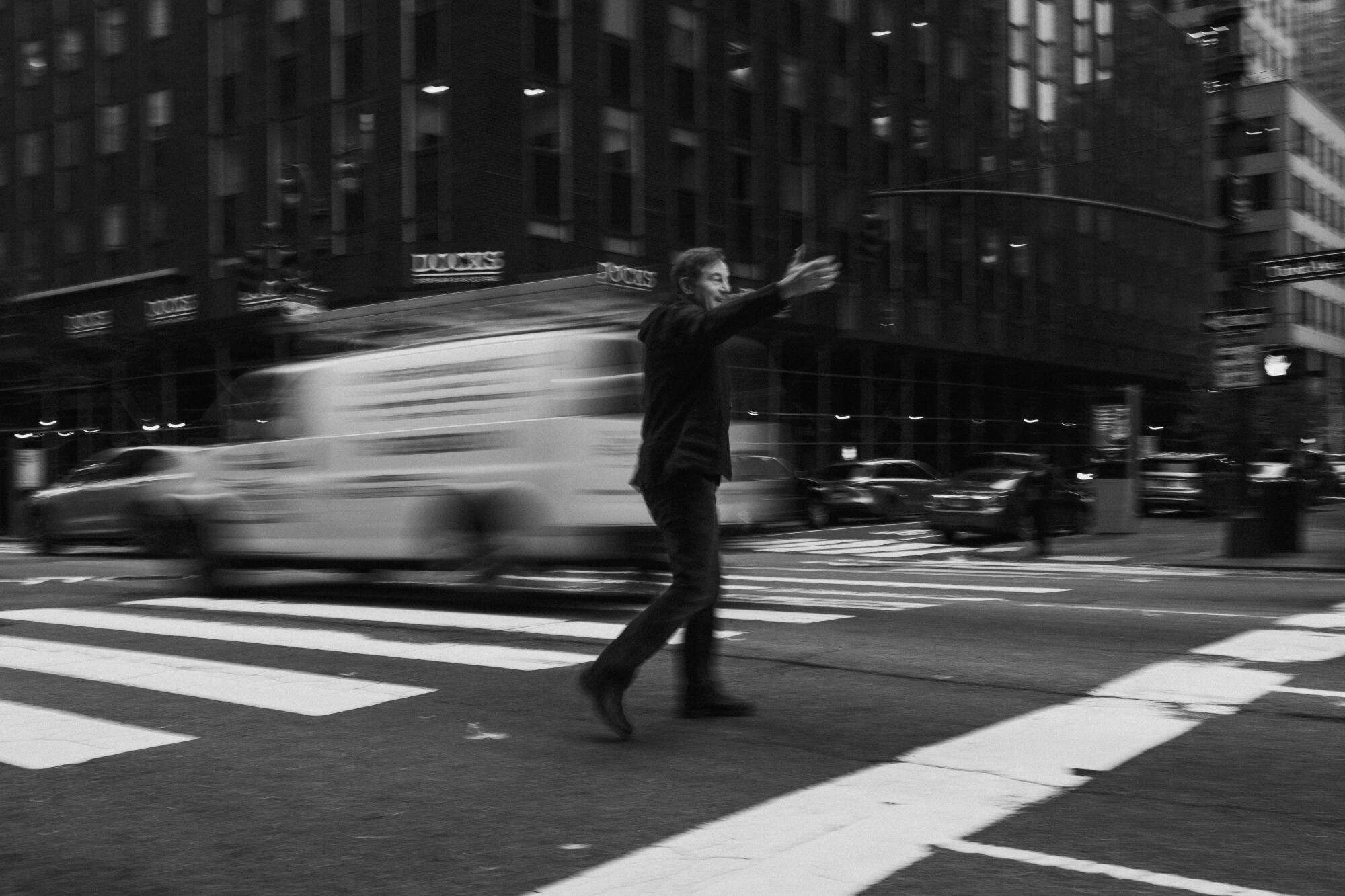 A black-and-white photo of a man standing in the crosswalk of a busy New York street as a car passes by.