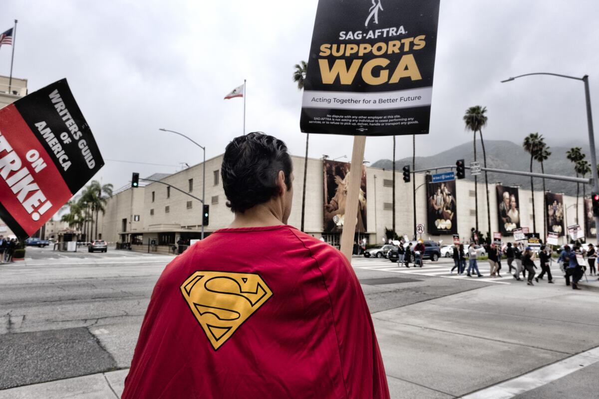 A picketer in a Superman costume holds a sign supporting WGA