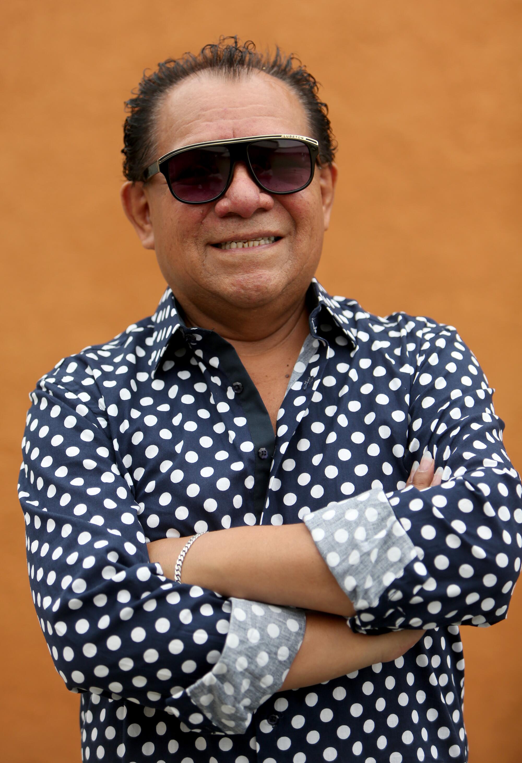 A man in a patterned button-down and black sunglasses crosses his arms 