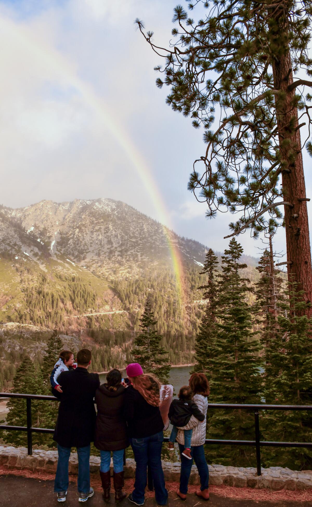 Four adults, two of them holding small children, stand at a railing with a view of mountains, ocean and a full rainbow.